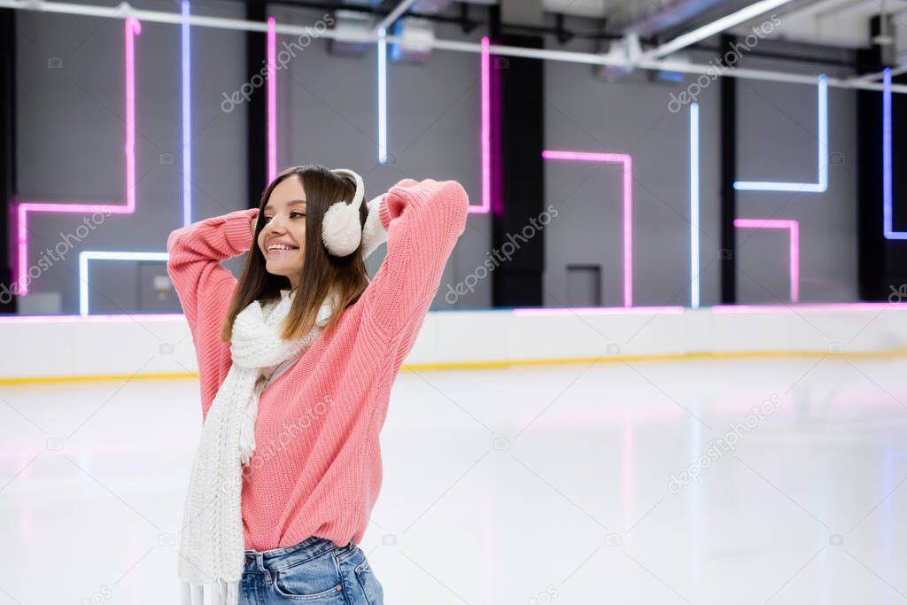 smiling young woman in pink sweater, scarf and ear muffs on ice rink 