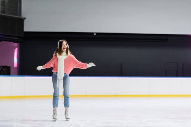 Beyaz eşarplı, kazaklı ve kulaklıklı buz pateni pistinde kayan hayretler içinde bir kadın.