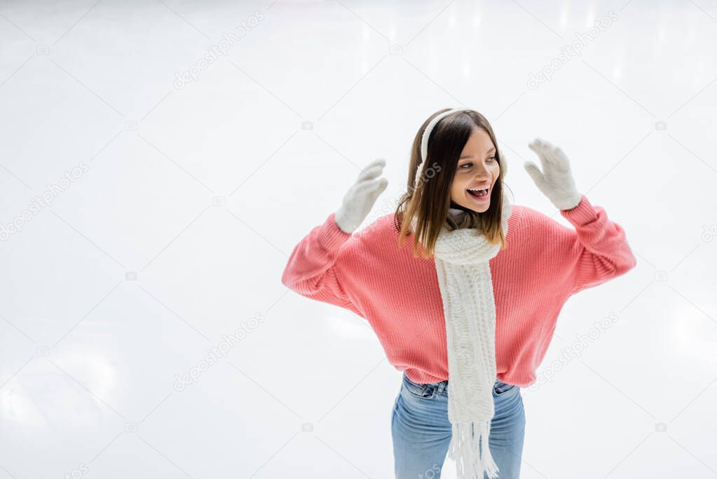 positive woman in pink sweater and white ear muffs gesturing on ice rink 