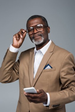 Gözlük ayarlayan akıllı telefonlu kıdemli Afro-Amerikan iş adamı gri kameraya bakıyor.