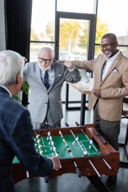 heyecan verici kıdemli işadamı, masa futbolu ve çok kültürlü meslektaşlarının yakınında zafer jesti yapıyor.