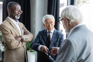 Kıdemli Asyalı işadamı, Afrika kökenli, kollarını kavuşturmuş Amerikalı bir adamın yanındaki bulanık iş ortağına evrak çantası veriyor.