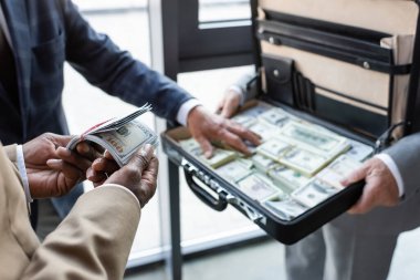Afrika kökenli Amerikalı iş adamının, bulanık meslektaşlarının yanında dolar banknotları ve para dolu evrak çantası tutarken çekilmiş görüntüsü.