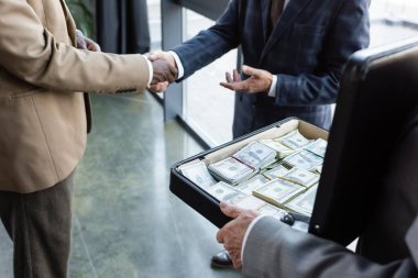 partial view of interracial business colleagues shaking hands near man holding briefcase with money clipart