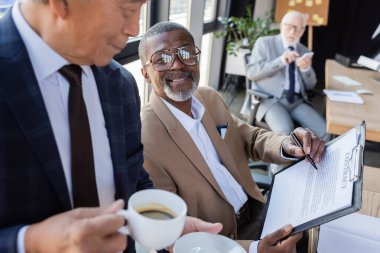 Afrika kökenli Amerikalı meslektaşının yanında bir fincan kahveyle sözleşmeye işaret eden kıdemli Asyalı iş adamı.