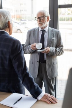 Gözlüklü kıdemli işadamı bulanık meslektaşıyla konuşurken elinde bir fincan kahve tutuyor.