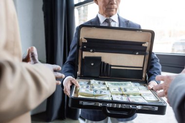 Bulanık iş ortaklarının yanında dolar banknotları olan bir evrak çantası taşıyan kıdemli adamın görüntüsü.