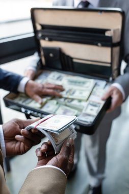 Afrika kökenli Amerikalı iş adamının bulanık meslektaşlarının yanında dolar ve para dolu evrak çantası tutması.