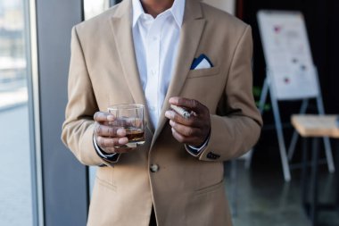 cropped view of senior african american businessman with cigar and glass of whiskey in office clipart