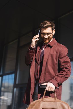 Trençkotlu gülümseyen iş adamı dışarıda akıllı telefondan konuşurken elinde çanta ve defter tutuyor. 