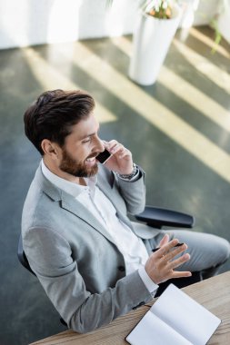 Ofiste defterin yanında akıllı telefondan konuşan neşeli iş adamının yüksek açılı görüntüsü. 