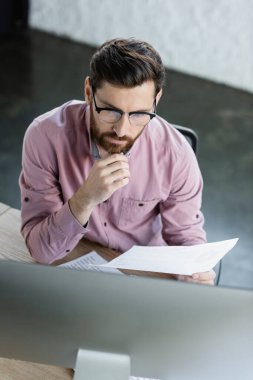 Ofiste bilgisayarın yanında tişörtlü bir işadamının kağıt tutarken yüksek açılı görüntüsü 