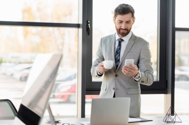 Ofiste dizüstü bilgisayar ve defterin yanında bir iş adamı elinde bardak ve cep telefonu tutuyor. 