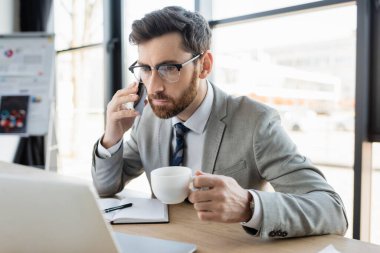Ofiste dizüstü bilgisayarın yanında akıllı telefondan konuşan bir işadamı. 