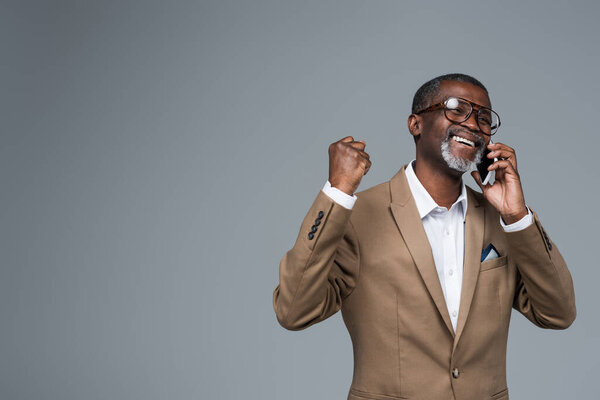 excited african american businessman showing success gesture while talking on cellphone isolated on grey