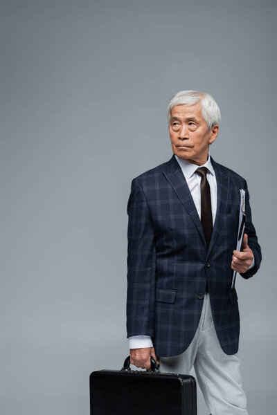 serious asian businessman with briefcase and clipboard looking away isolated on grey