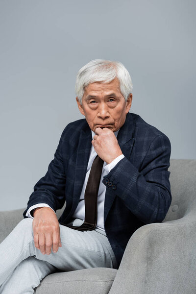 senior asian businessman in formal wear looking at camera while sitting in armchair isolated on grey