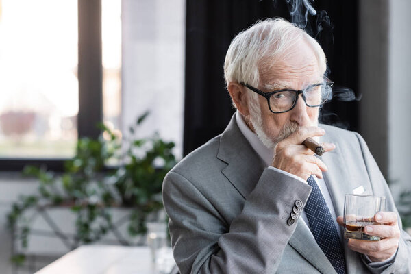 elderly businessman with glass of whiskey looking at camera while smoking cigar in office