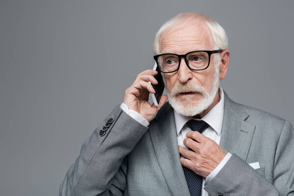 senior thoughtful businessman touching tie while talking on cellphone isolated on grey