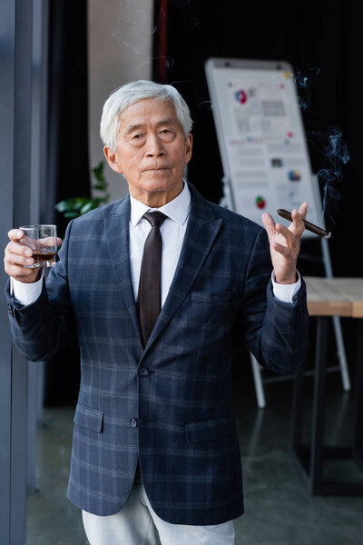 senior and confident asian businessman with cigar and glass of whiskey looking at camera in office