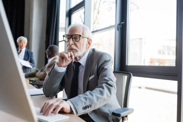 Idősebb Üzletember Szemüvegben Számítógépen Dolgozik Homályos Multikulturális Kollégák Közelében — Stock Fotó