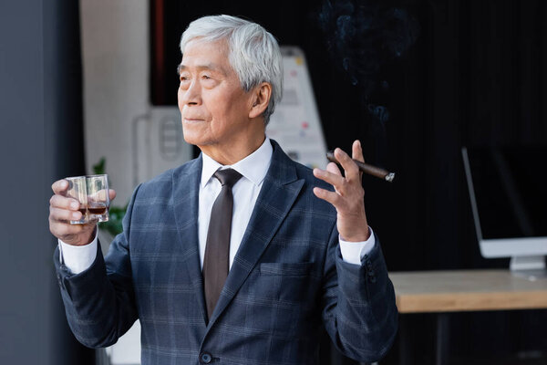 elderly asian businessman with cigar and glass of whiskey looking away in office
