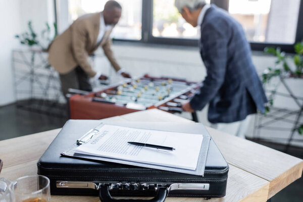 selective focus of contract on briefcase near senior businessmen playing table football on blurred background