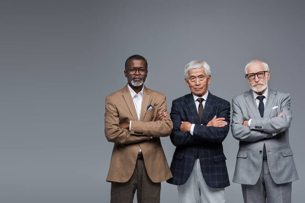 serious and confident multiethnic business partners standing with crossed arms isolated on grey
