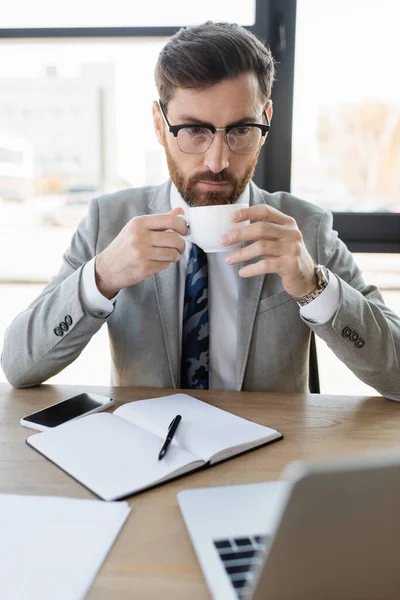 Biznesmen Trzymając Kubek Patrząc Laptopa Pobliżu Notebooka Biurze — Zdjęcie stockowe