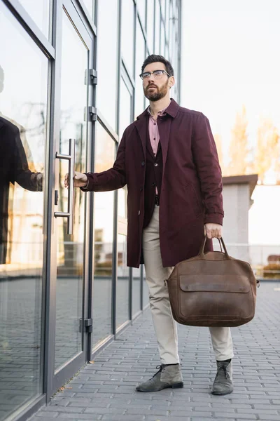 Businessman in trench coat holding bag near door of building outdoors