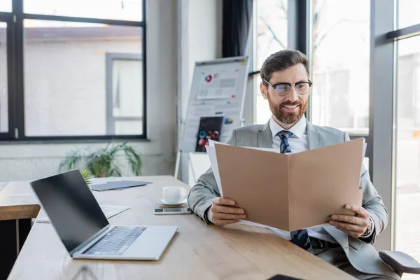 Glimlachende Zakenman Zoek Naar Papieren Map Buurt Van Laptop Kantoor — Stockfoto