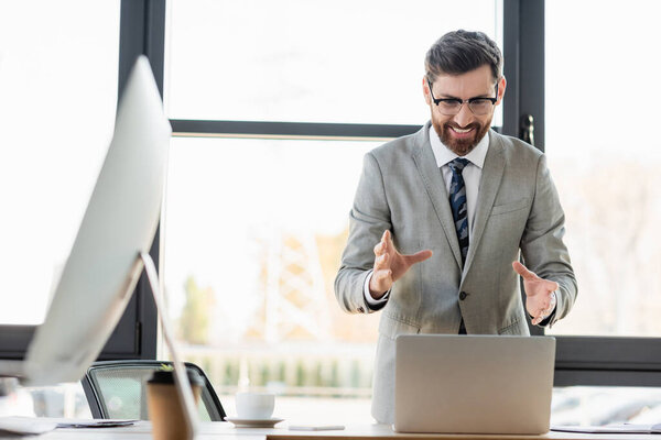 Cheerful businessman having video chat on laptop near coffee in office 