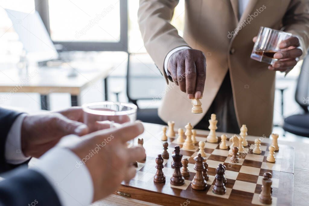 cropped view of african american businessman with glass of whiskey playing chess with blurred colleague