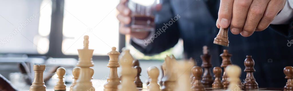 cropped view of businessman with blurred glass of whiskey playing chess in office, banner