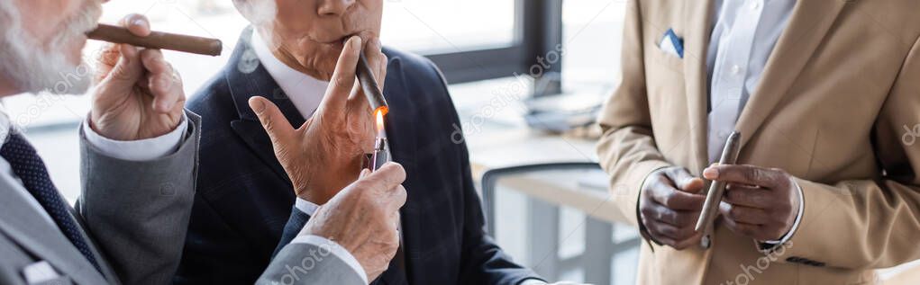 cropped view of senior businessman lighting cigar of business partner near blurred african american colleague, banner
