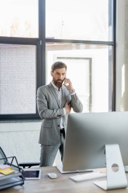 İş adamı akıllı telefondan konuşuyor ve ofisindeki bilgisayar monitörüne bakıyor. 