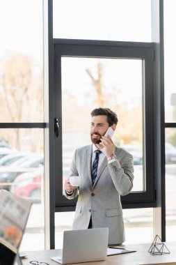 Pozitif iş adamı akıllı telefonla konuşuyor ve ofisinde kahve fincanı tutuyor. 
