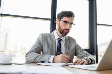 Gözlüklü bir iş adamı dizüstü bilgisayarın yanında ve ofisinde yazıyor. 