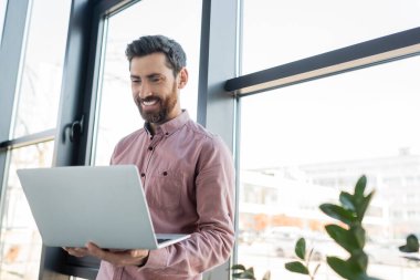 Ofisteki pencerenin yanında dizüstü bilgisayar kullanan gömlekli mutlu bir işadamı. 