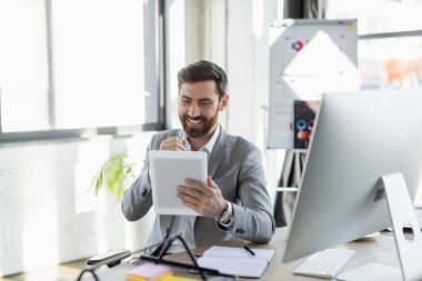 Ofiste bilgisayarın ve defterin yanında dijital tablet kullanarak gülümseyen takım elbiseli işadamı 