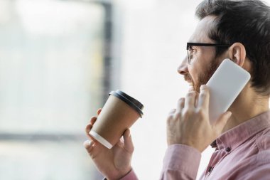 Pozitif iş adamının akıllı telefonla konuşmasının ve ofisinde kağıt bardak tutmasının yan görüntüsü 