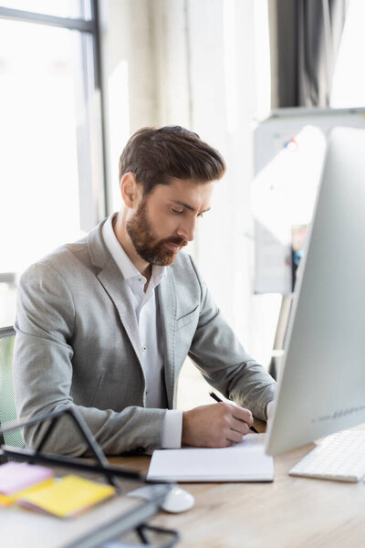 Bearded businessman writing on notebook near computer in office 