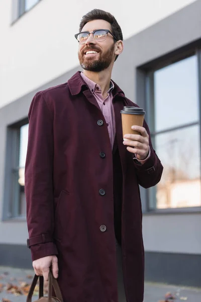 Bearded businessman in trench coat holding bag and paper cup on urban street
