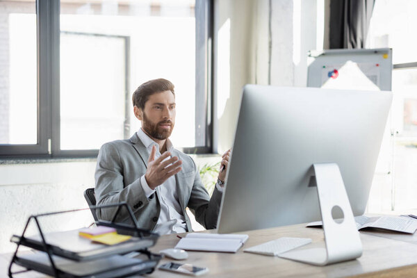 Bearded businessman having video call on computer near newspaper and notebook in office 