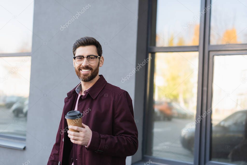 Businessman in trench coat smiling at camera while holding coffee to go on urban street 