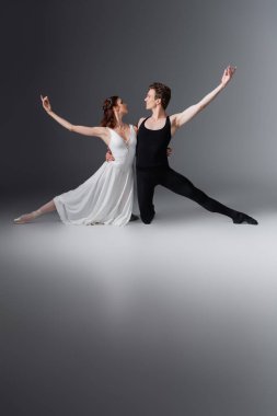 full length of graceful ballerina in white dress dancing with partner looking at each other on dark grey clipart