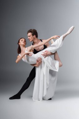 Koyu gri üzerinde dans ederken beyaz elbiseli zarif balerini destekleyen uzun boylu bir adam.