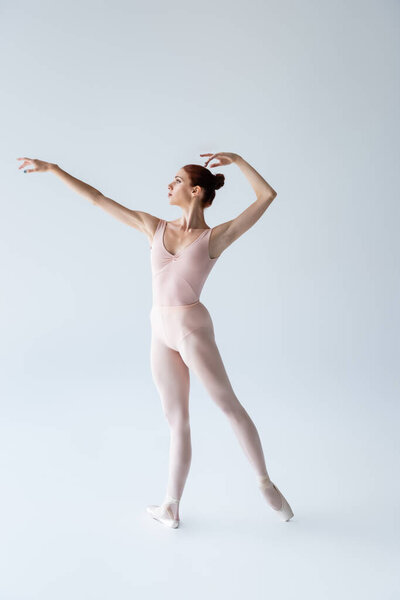 full length of young ballerina in bodysuit gesturing while standing on grey
