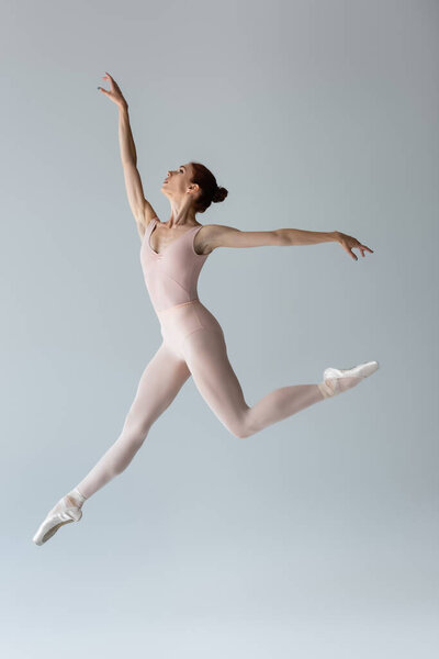 full length of graceful ballerina in bodysuit levitating and gesturing on grey