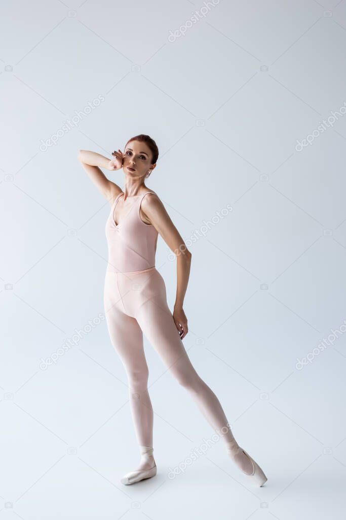full length of elegant ballerina in bodysuit looking at camera on grey